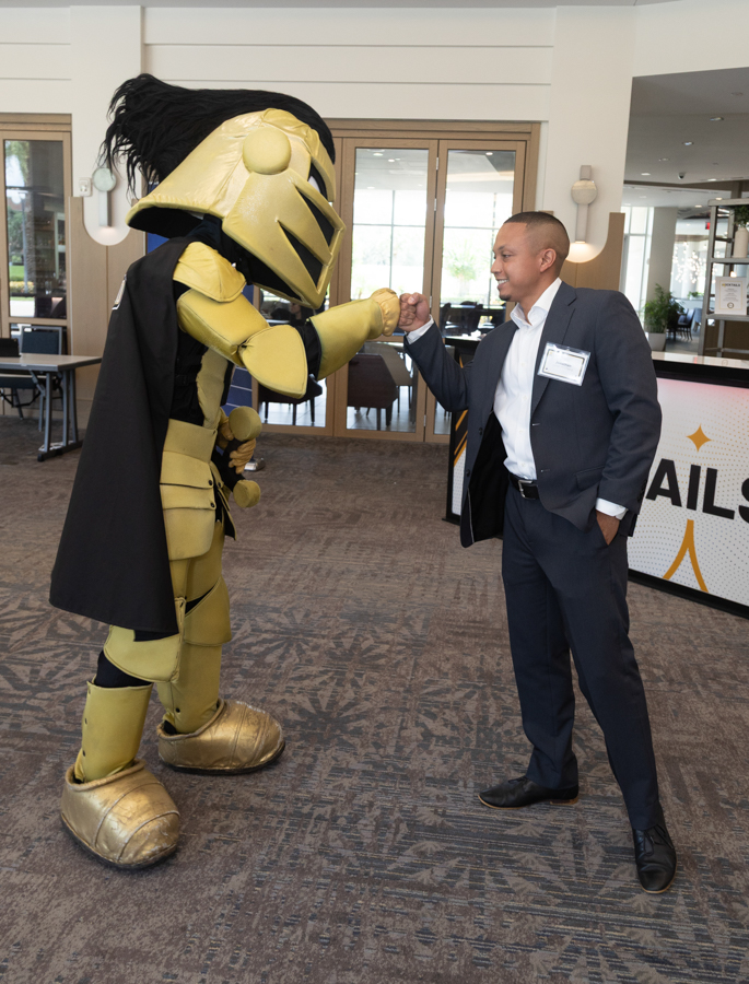 Man in a suit fist-bumps a large gold knight mascot at a corporate event in a bright lobby backdrop.