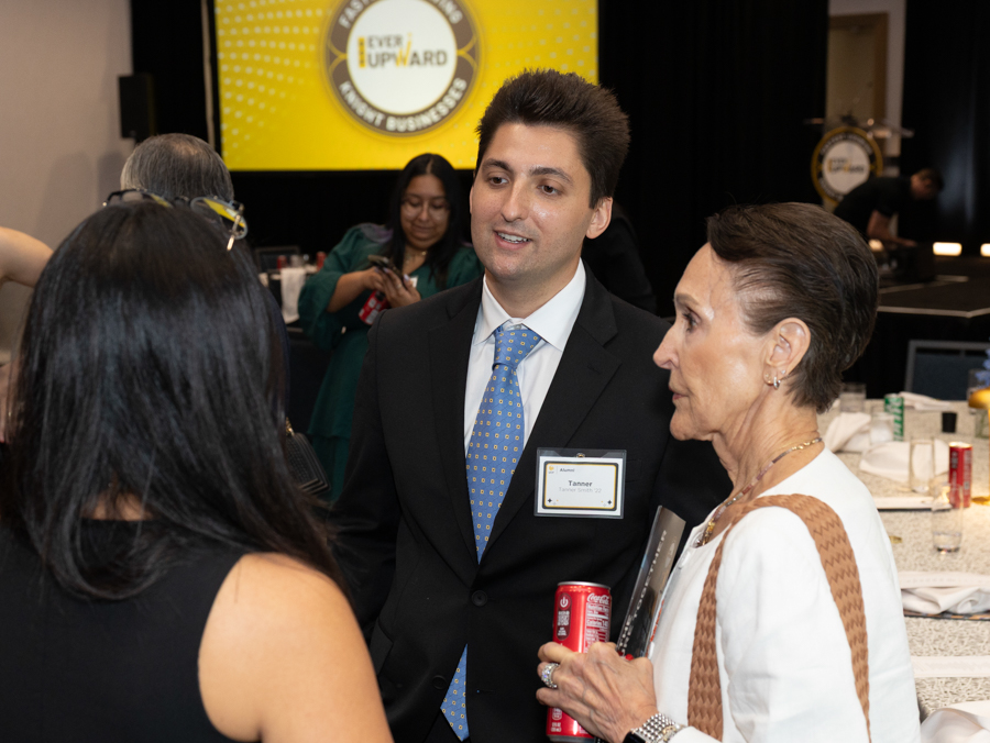 Man in a suit with a name badge chats with attendees at a conference reception, yellow stage backdrop behind him.