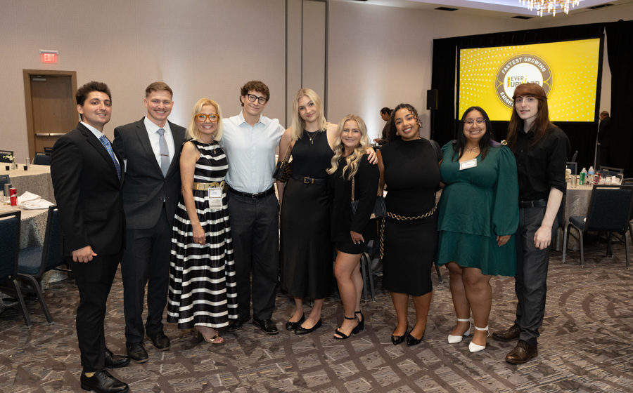 Nine professionals in formal attire pose for a group photo at a conference banquet room.