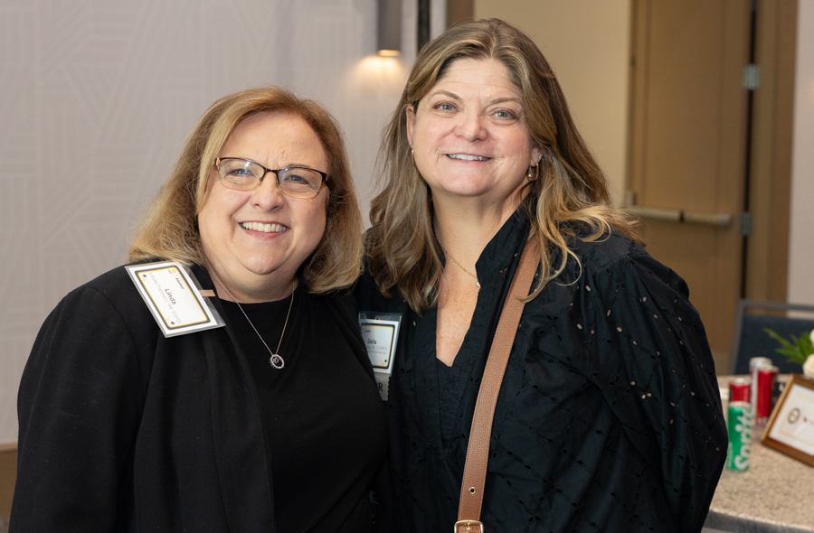 Two smiling women with name badges posing together at an event. One wears glasses and a black top; the other has a brown bag strap over a dark blouse.