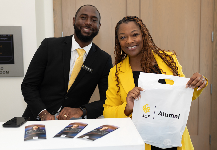 Smiling man in a black suit and yellow tie stands beside a smiling woman in a yellow blazer holding a UCF Alumni bag at a registration desk with brochures on the table.