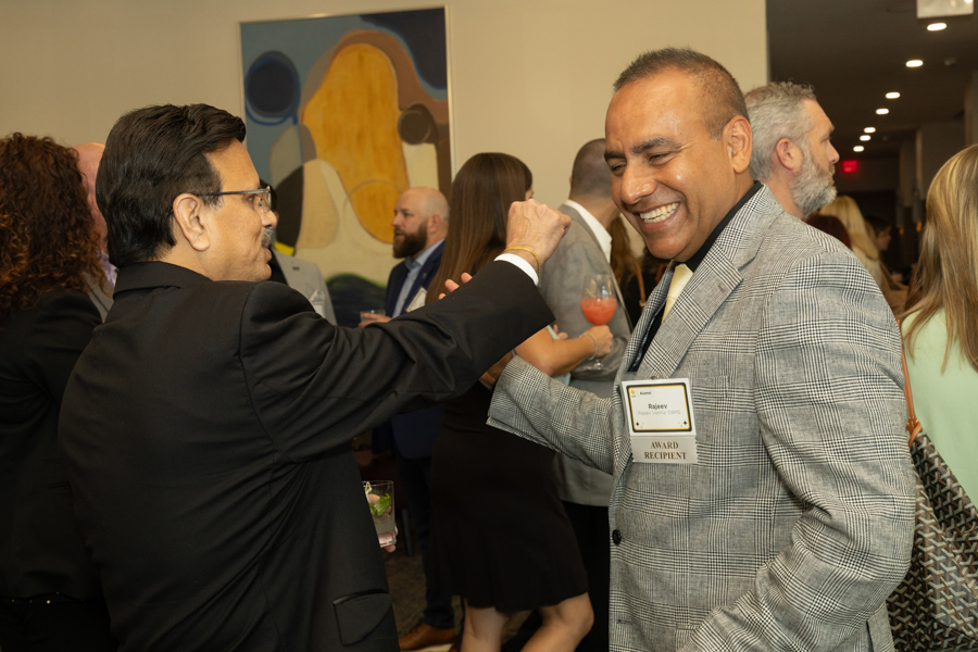 Two men in a crowd share a fist bump at an awards reception, smiling and congratulated by colleagues in the background.