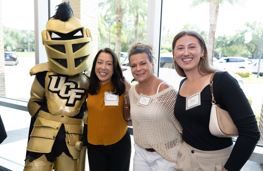 Four people posing with a gold UCF knight mascot by a window at an event, smiling for the camera.