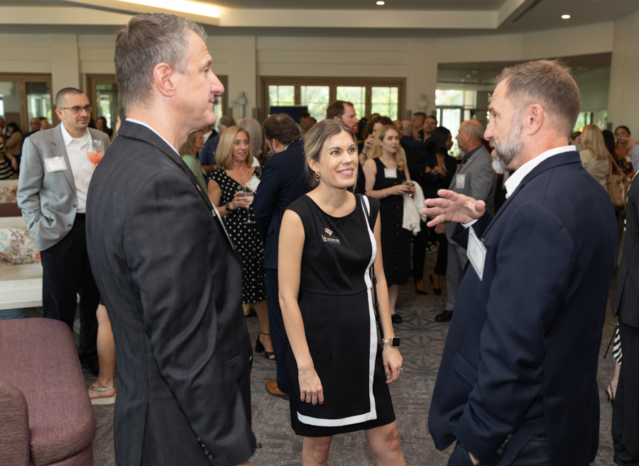 Professionals networking at a bright indoor event; a woman in a black dress chats with two men in suits as others mingle in the background.