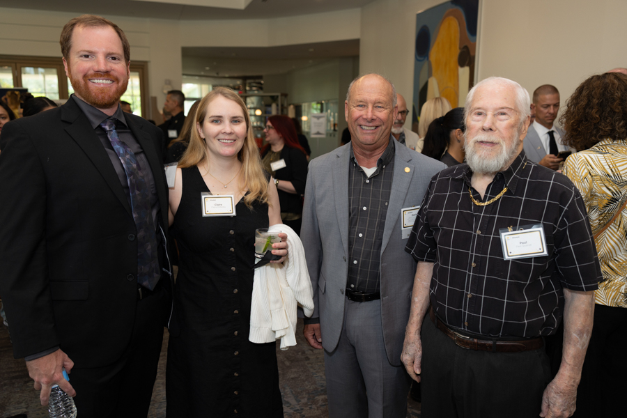 Group of four adults posing and smiling at a social event, name badges visible on their chests. In a bright, modern room with art on the wall.