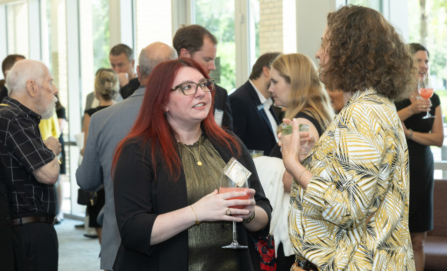 Two women converse at a networking reception, holding drinks, with other guests mingling in the background in a bright venue.