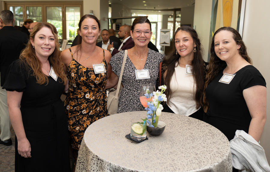 Five women in dresses stand around a high round table with a floral centerpiece, smiling at the camera at a reception for networking.