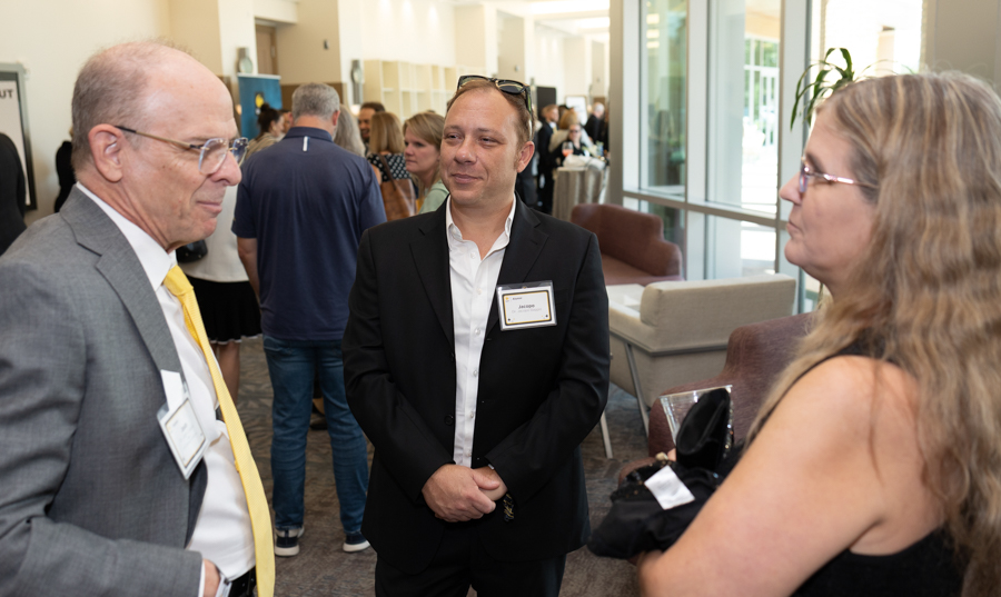 Three professionals converse at a networking event in a bright lobby; man in a black suit smiles at center, an older man in gray with a yellow tie on left, woman with long hair on right holds a bag and badge visible.