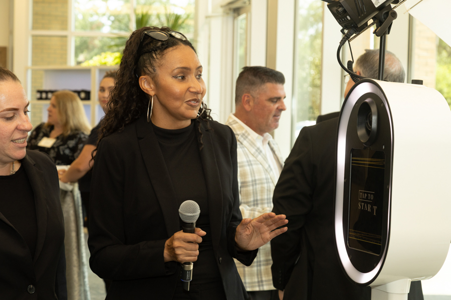 Woman in a black blazer with a microphone speaks at a tech event, standing beside a white circular touchscreen kiosk with 'Tap to Start' on the screen cleared for interaction.