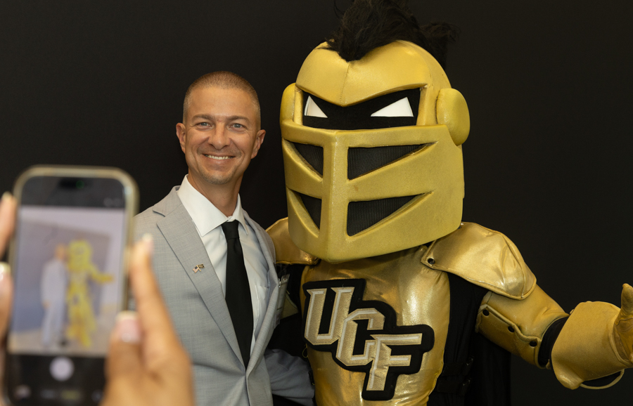 Smiling man in a light gray suit posing with a large gold knight mascot in front of a dark background.