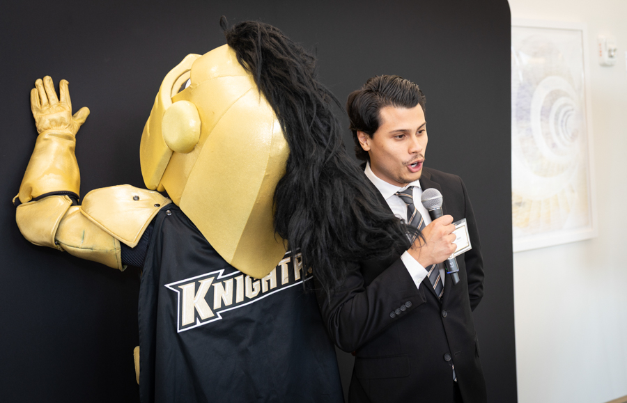 Man in a suit speaks into a handheld microphone beside a large gold knight mascot with a 'Knights' logo on its cape against a dark backdrop.