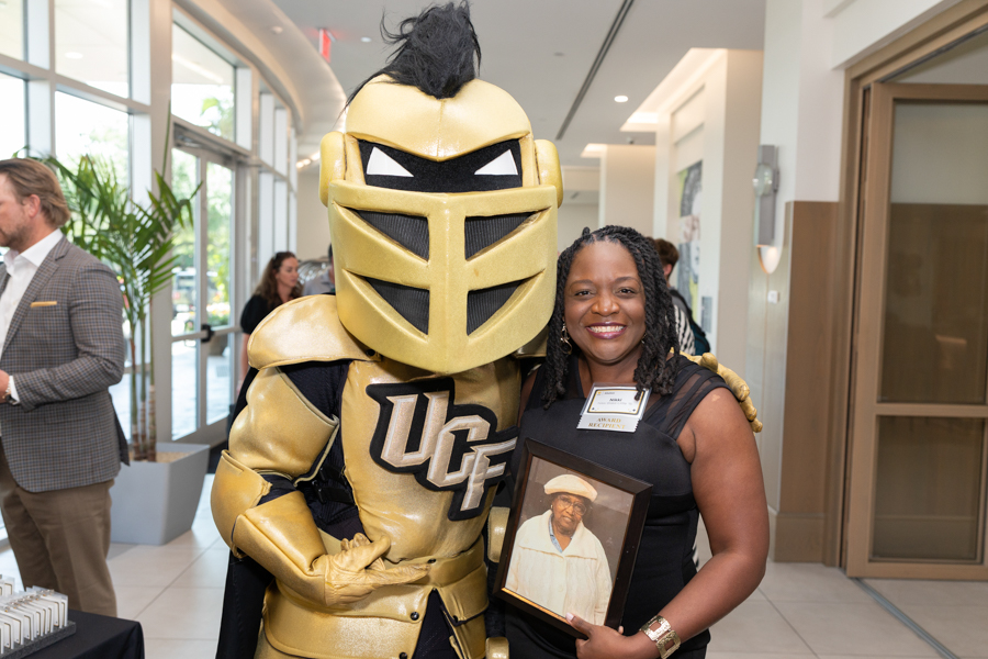 Golden Knight mascot in shiny gold armor poses with a smiling woman at an event; she holds a framed photo of a man in a white hat.