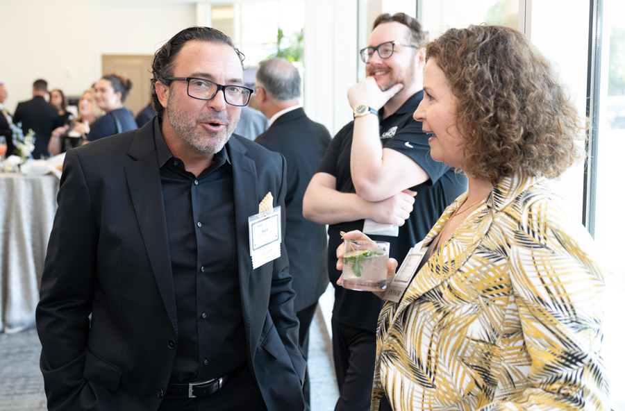 Two professionals chat near a window at a networking event; a man in a black suit and a woman in a yellow patterned blazer hold drinks and wear name badges.