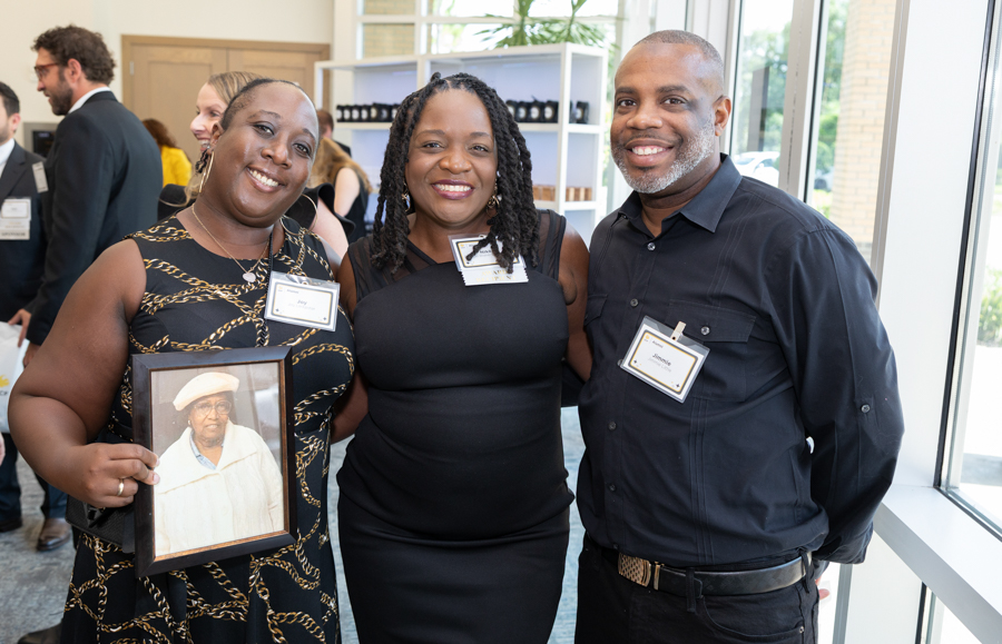 Three adults posing for a photo at a networking event; the woman on the left holds a framed portrait of an elderly person, all smiling at the camera.