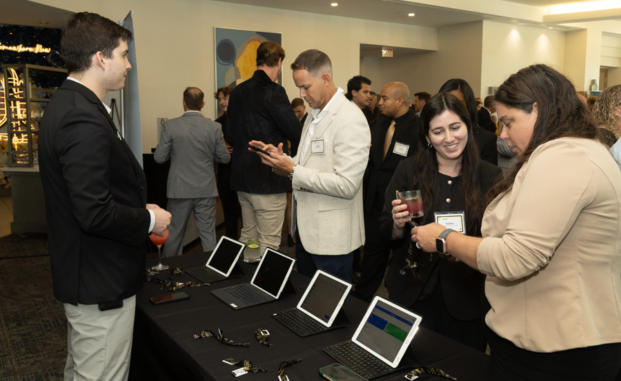 Networking event with attendees around a table displaying multiple laptops and tablets for demos, drinks in hand.