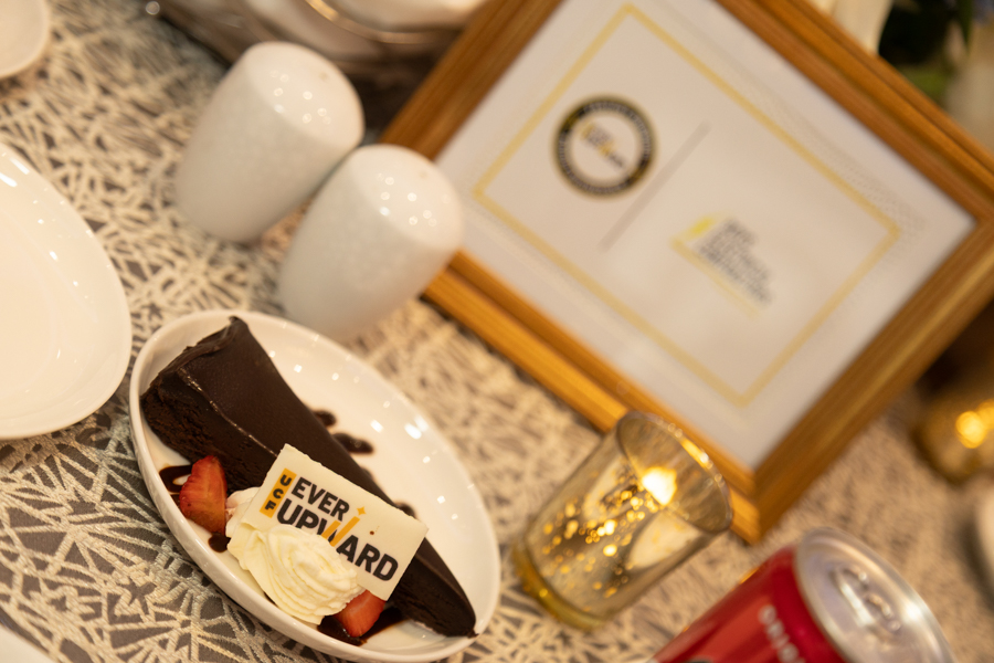 Chocolate cake slice with whipped cream and strawberries on a plate, topped by a ‘Never Give Up’ card; framed certificate and decorative candle holder in the background.