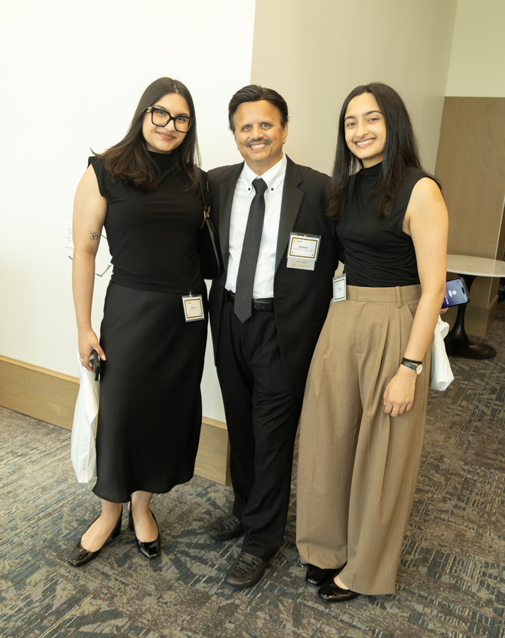 Three professionally dressed people posing together in an office/conference area, two women on either side of a man, all wearing name badges and smiling at the camera.