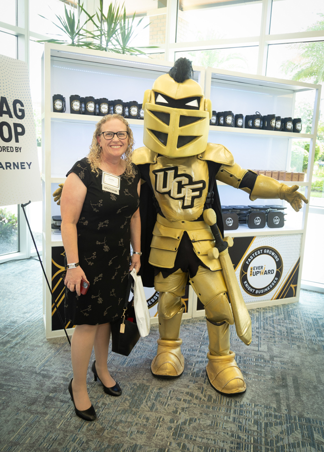 Smiling woman in a black dress posing with a large gold knight mascot at a conference booth.