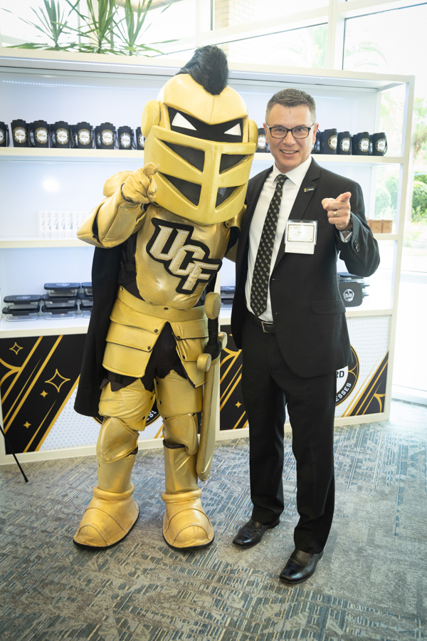 Gold knight mascot in armor posing with a man in a suit, pointing at the camera at a promotional event.