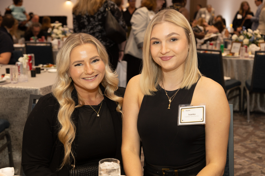 Two smiling women in black dresses sit at a banquet table during a formal event.