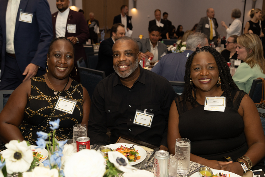 Three smiling guests wearing name badges sit at a round banquet table with dinner plates and drinks on a formal event.