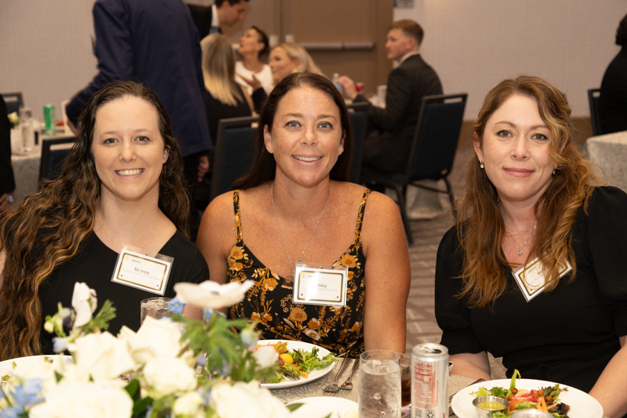 Three women sit at a banquet table, smiling for the camera, each wearing a name tag during a formal dinner event.