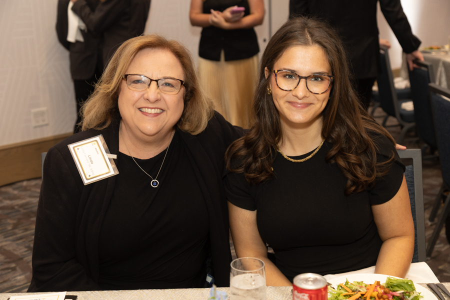 Two smiling women sit at a banquet table together, wearing black outfits and glasses; the woman on the left has a name badge on her lapel.