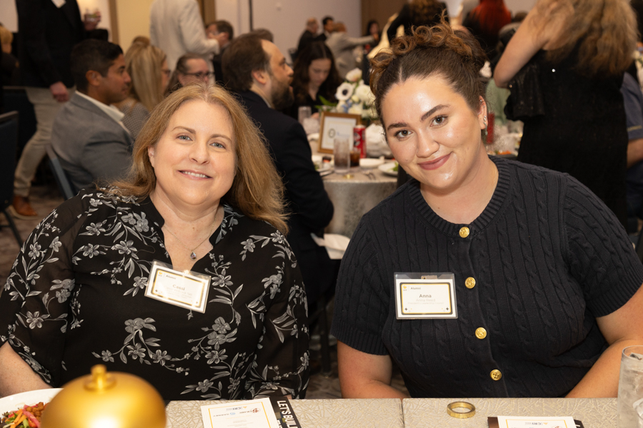Two women seated at a banquet table, smiling for the camera at a formal event, each wearing a name badge.
