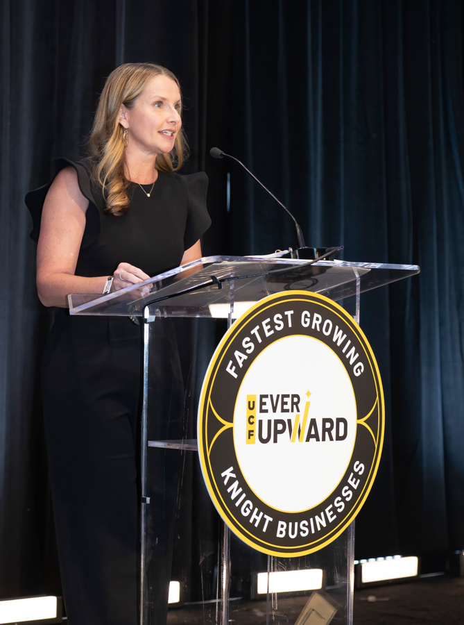 Speaker in a black dress at a clear podium during a formal event, with a circular Ever Upward logo on the podium seal.