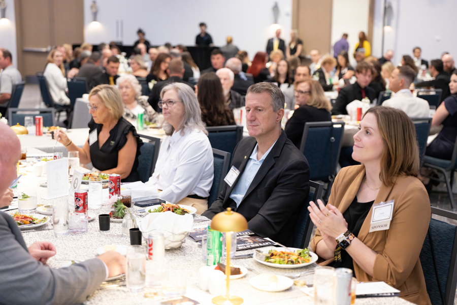 People in business attire dining at banquet tables during a conference event, with food and drinks on the table.