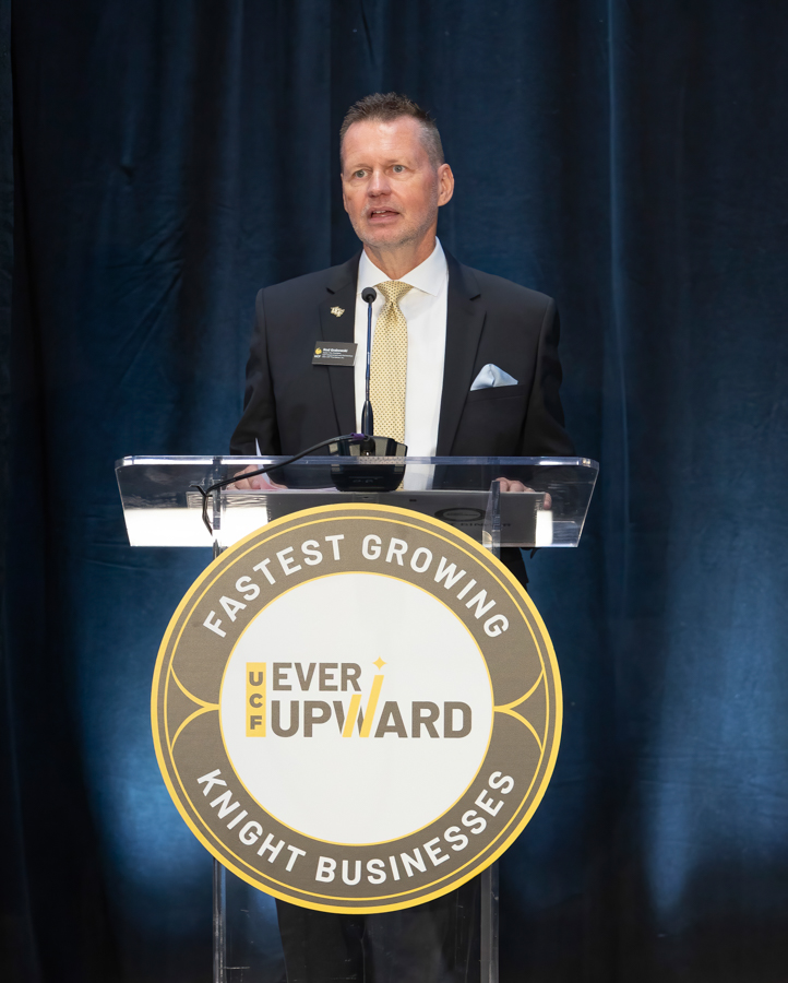 Man in a dark suit and yellow tie speaks at a clear podium with a circular event sign reading 'Fastest Growing Knight Businesses' and 'UPWARD' in the center.