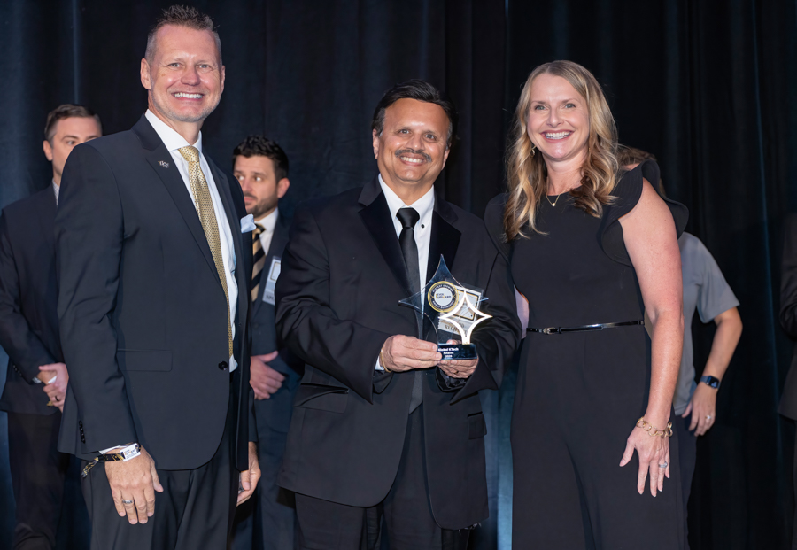 Center man in a black suit holding a star-shaped trophy on stage, flanked by a man in a suit and a smiling woman in a black dress.