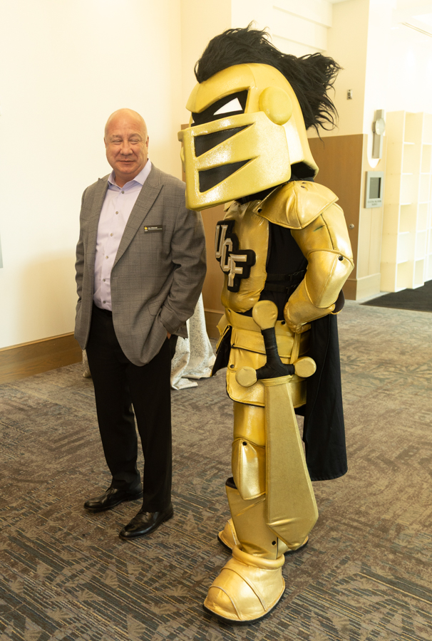 Man in a gray suit posing next to a gold knight mascot at an indoor event.