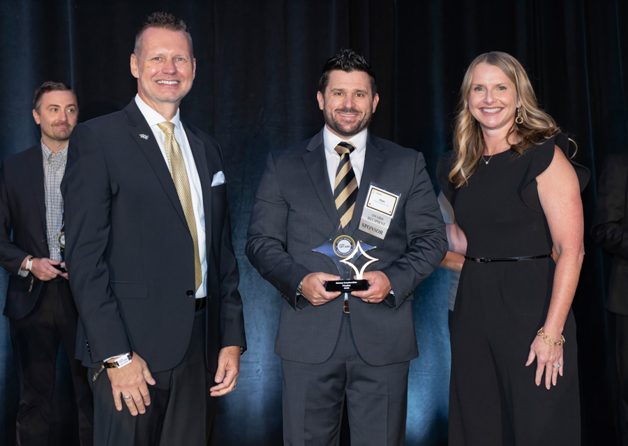 Three adults in formal attire on stage at an awards ceremony; center man holds a trophy, flanked by two colleagues smiling at the camera.