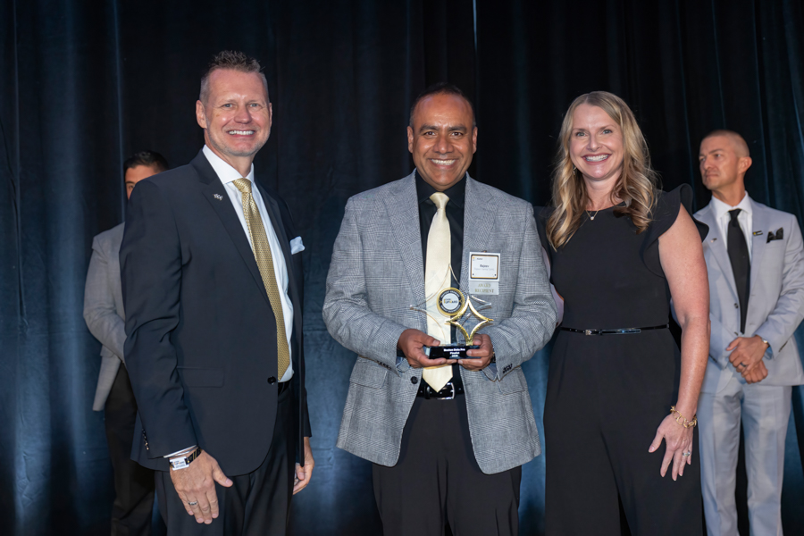 Man in gray blazer holds an award on stage with two smiling colleagues nearby, black backdrop behind them.