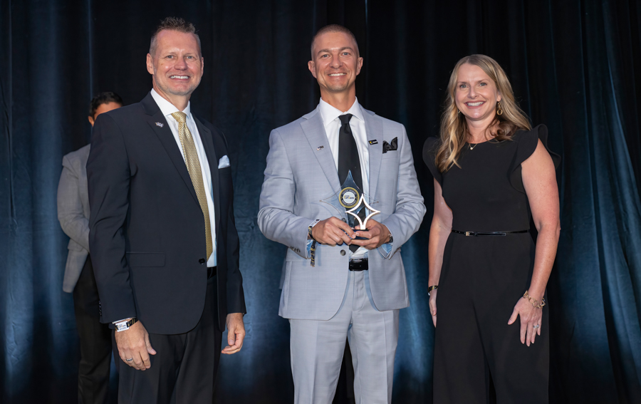 Three adults in formal attire on stage; center man in a light gray suit holds a star-shaped award while flanked by colleagues on a dark backdrop stage.