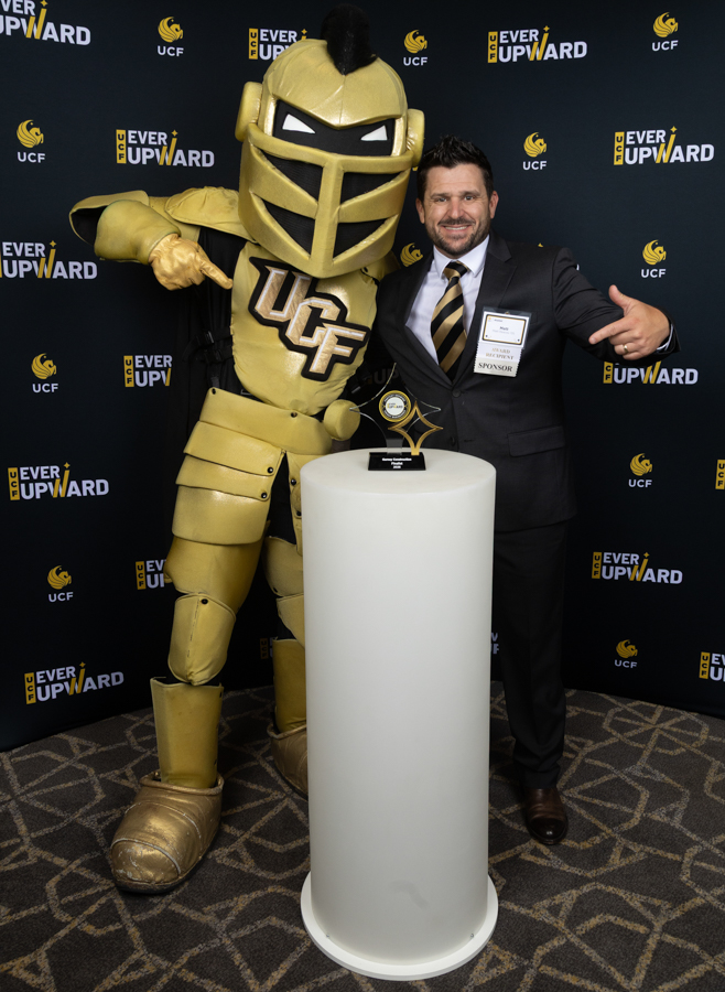 Man in a suit posing with a gold UCF knight mascot beside a trophy on a white pedestal at a UCF 'Ever Upward' event.