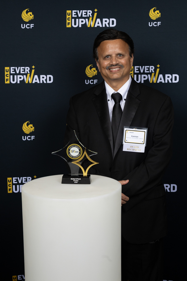 Smiling man in a black suit poses with a star-shaped award on a white pedestal at a UCF 'Ever Upward' event backdrop.