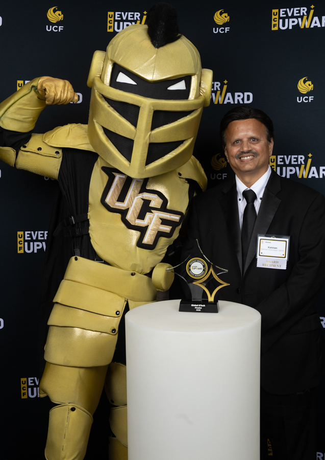 Gold knight mascot in UCF armor poses with a man in a suit at an awards event, trophy on a white pedestal in front.