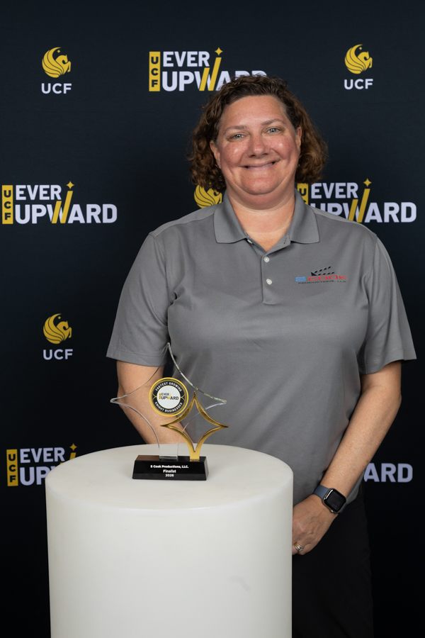 Woman in a gray polo stands beside a white pedestal with a trophy at a UCF Ever Upward event backdrop.