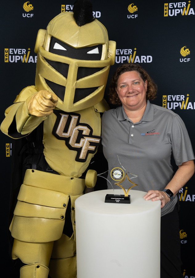 UCF knight mascot in gold armor posing with a smiling woman at an awards ceremony, a trophy on a pedestal nearby.