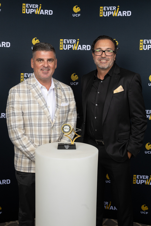 Two men in suits pose beside a white pedestal holding a trophy, with a dark backdrop featuring 'Ever Upward' and UCF logos.