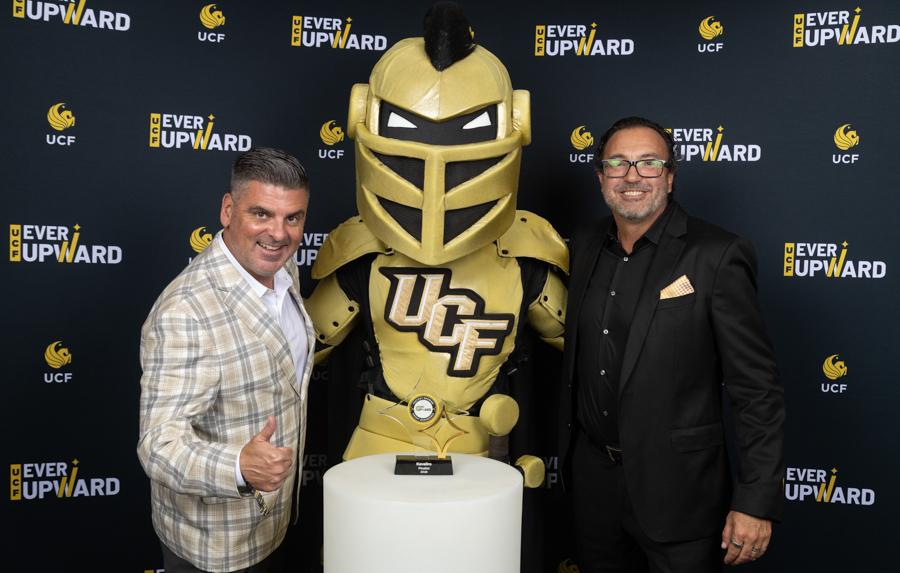 Two men in suits pose with a gold UCF Knight mascot and trophy on a pedestal against a UCF 'Ever Upward' backdrop.