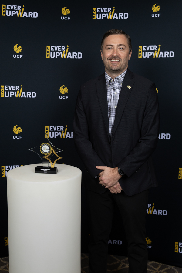 Smiling man in a dark suit and checkered shirt stands beside a white pedestal holding a glass star-shaped trophy, at an UCF 'Ever Upward' event backdrop.