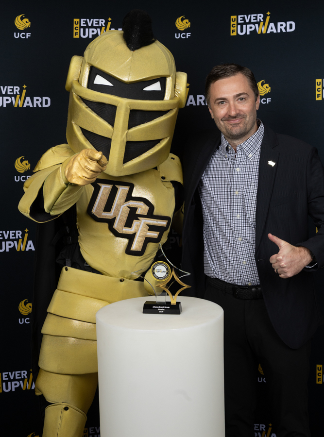 Man in a checkered shirt and blazer stands beside a gold UCF knight mascot, posing with a trophy on a pedestal at an event backdrop.
