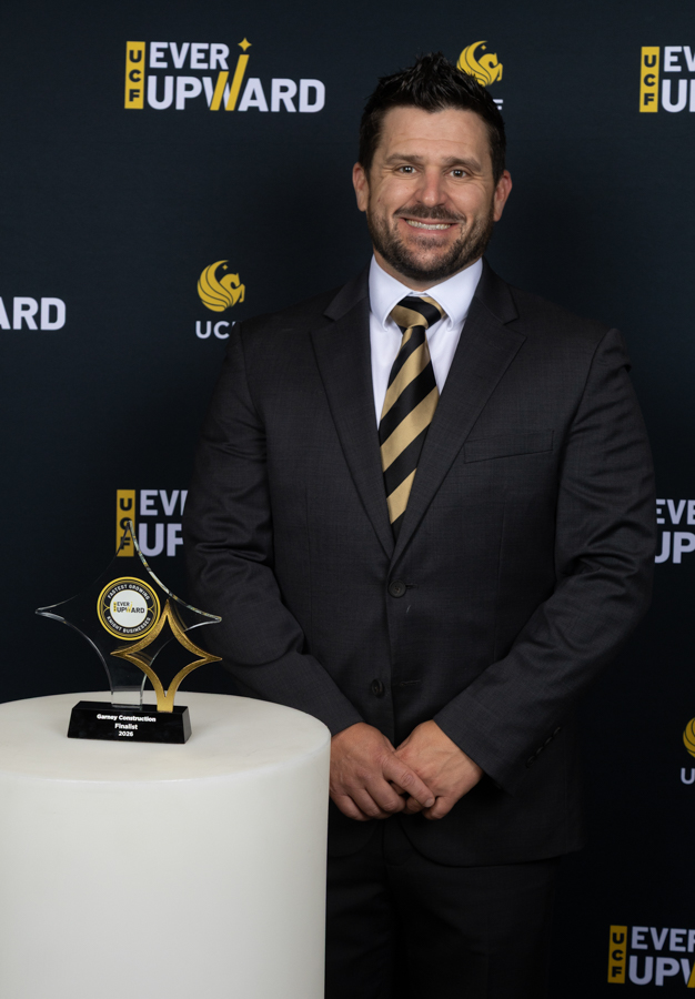 Man in a dark suit and striped gold-and-black tie standing beside a glass award on a white pedestal at an awards event backdrop.