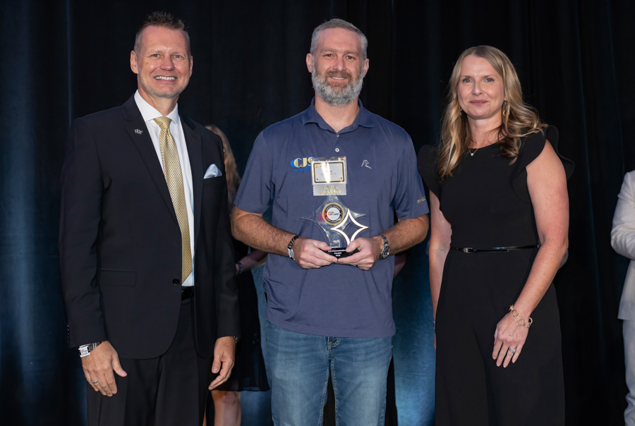 Man in a blue polo holding an award, flanked by a suited man and a woman in a black dress on stage.