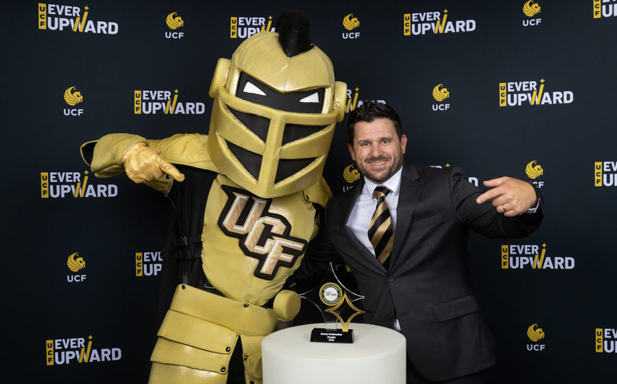 Man in a suit posing with a gold armored UFC mascot at the Ever Upward event, a trophy on a white pedestal in front of them.