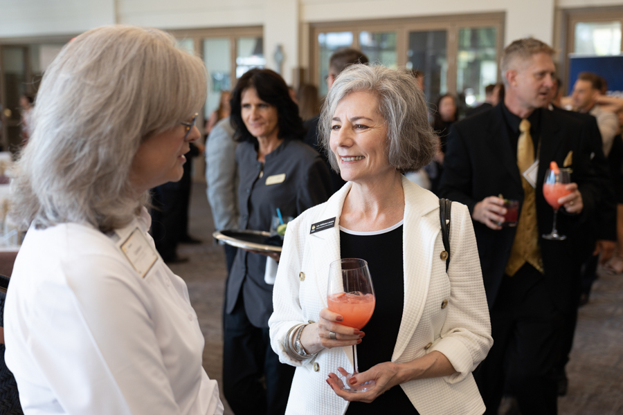 Two women mingle at a reception, one holding a pink cocktail and smiling as they chat.