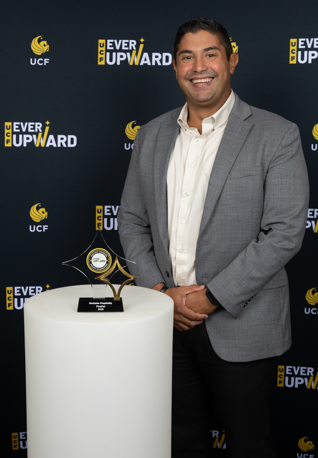 Man in a gray blazer and cream shirt poses beside a white pedestal with a clear star-shaped award; backdrop shows UCF 'Ever Upward' logos.